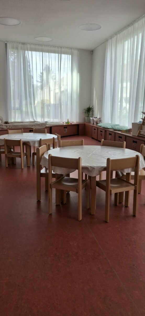 Třída s kulatými stoly s ubrusy a dětskými židličkami u prosklené stěny
